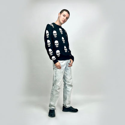Person wearing a black sweater with white skull patterns and light gray pants on a white background